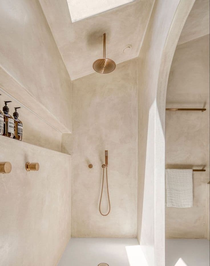 Waterproof Microcement Flooring in Jeddah Bathrooms Warm beige textured seamless Microcement Flooring and walls in a minimalist Jeddah bathroom shower area with brass fixtures and a skylight, showing a seamless Microcement Flooring installation by Floroz KSA.