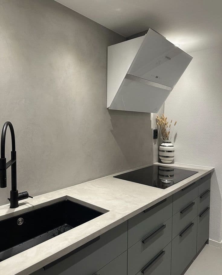 Easy-Clean Satin Microcement for Kitchens & Countertops Modern kitchen featuring grey microcement splashback and countertop with a black sink, induction cooktop, and white extractor hood.
